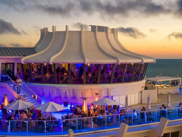 An exterior view of a multi-level building with people dining under umbrellas, illuminated with blue lights, set against a sunset over the ocean.
