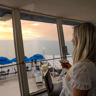 A woman with long blonde hair is holding a coffee cup, gazing out at a sunset over a beach with blue umbrellas, viewed from a window.