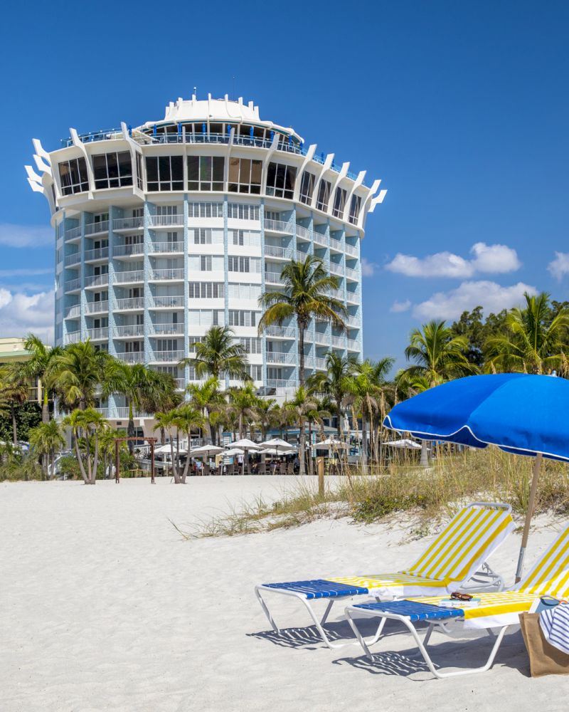 A beach scene with two lounge chairs under a blue umbrella, a tall building in the background, and palm trees under a blue sky with clouds.