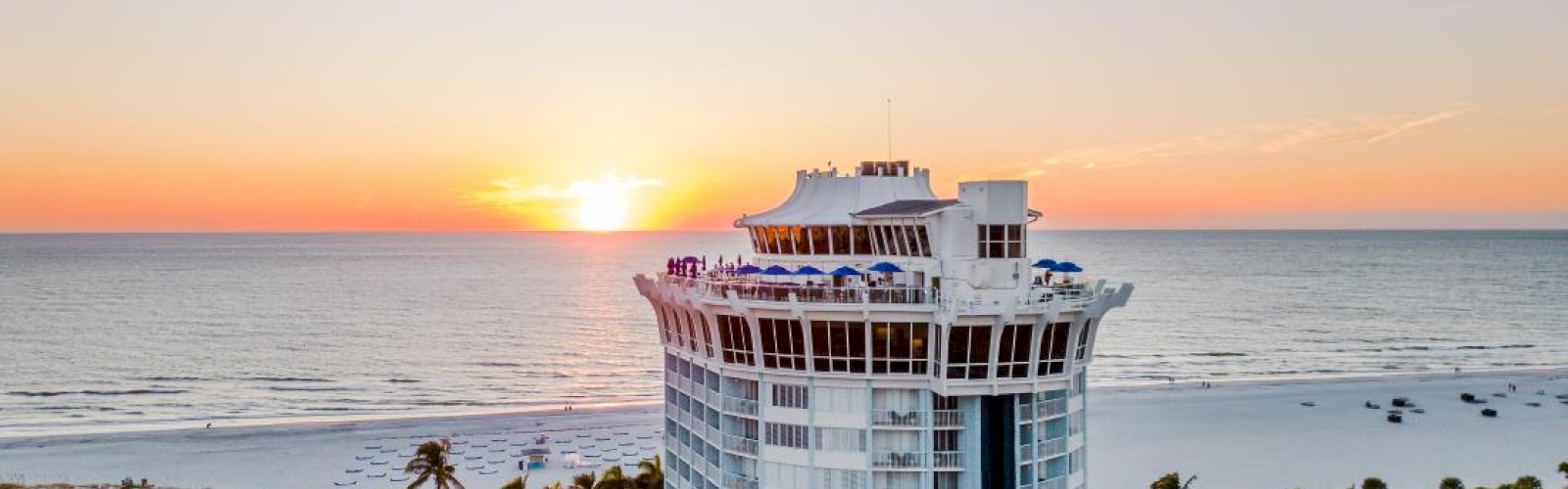 A tall, circular hotel building by the beach with a sunset view, surrounded by palm trees and other structures.
