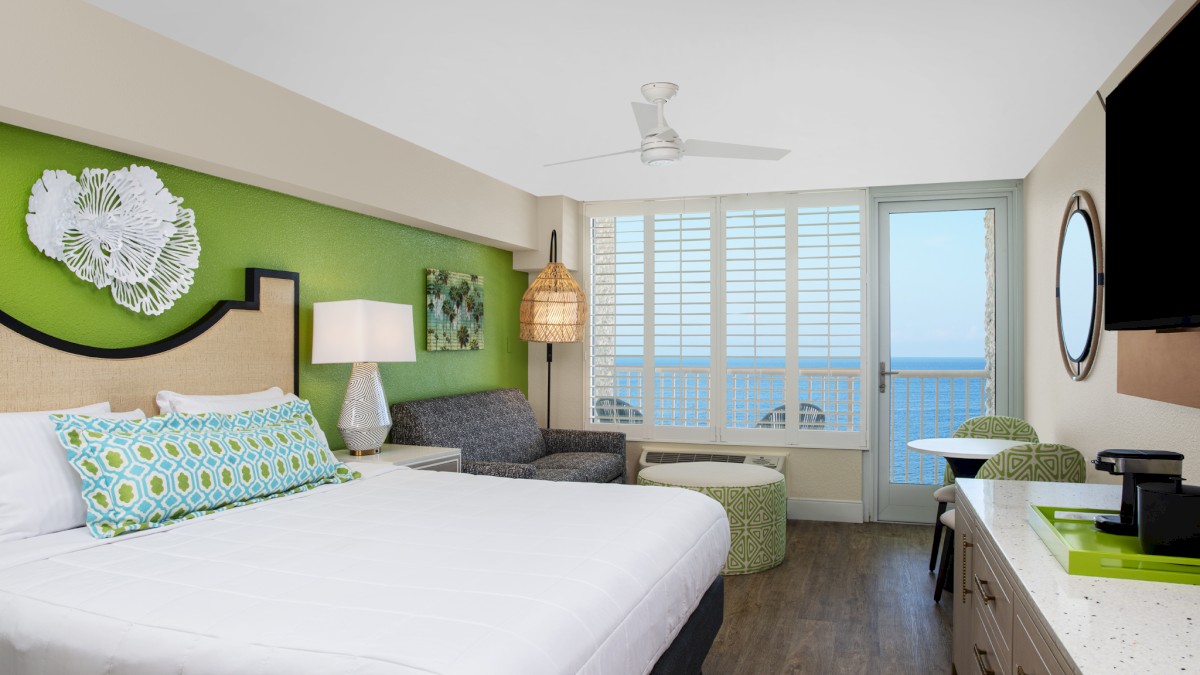 This image shows a modern hotel room with a bed, sofa, desk, and a TV, decorated with green accents and offering an ocean view through large windows.