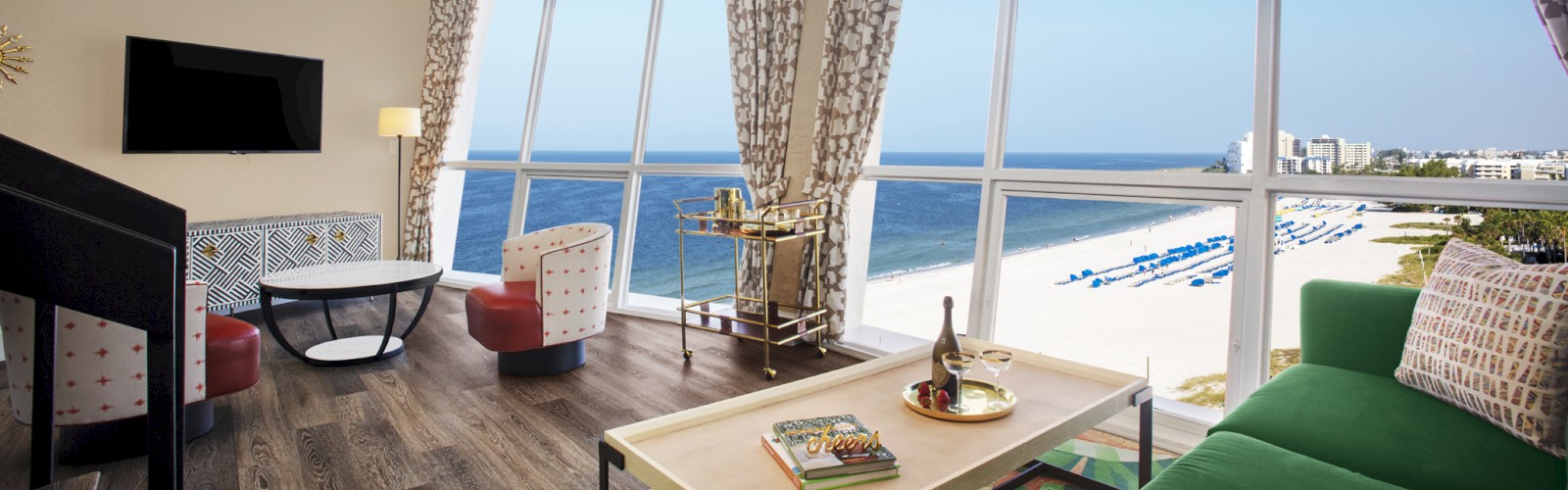 The image shows a bright, modern room with large windows overlooking a beach. The room has comfortable seating, a coffee table, and a wall-mounted TV.