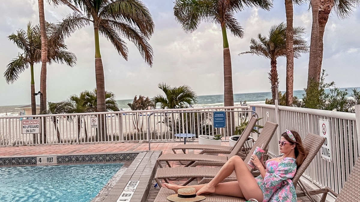 A person lounges by a pool on a sunny day, surrounded by palm trees, a pink bag beside them, and they appear to be using a smartphone.