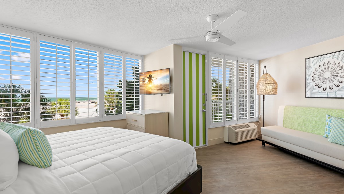 A bright bedroom with large windows, a bed, a couch, a ceiling fan, an air conditioner, and a wall-mounted TV displaying a beach scene ends the sentence.