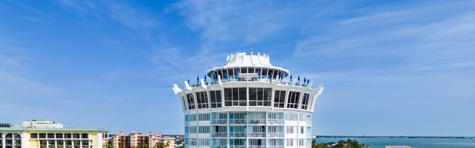 A tall, round hotel surrounded by palm trees and other buildings, with a view of the coastline and ocean in the background, under a blue sky.