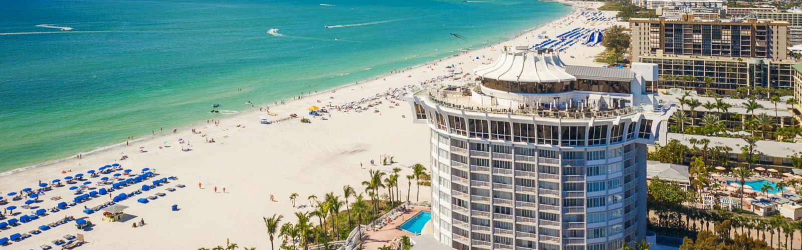 Beach with white sand, turquoise waters, beach chairs with blue umbrellas, and a round, multi-story building. Other buildings and palm trees in view.
