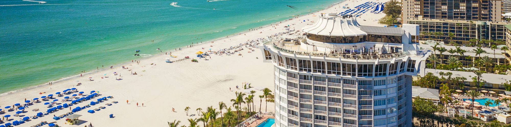 Beach with white sand, turquoise waters, beach chairs with blue umbrellas, and a round, multi-story building. Other buildings and palm trees in view.