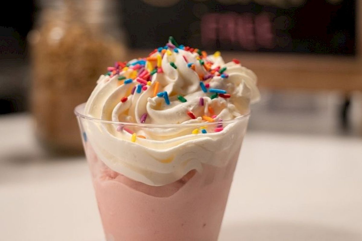 The image shows a cup of soft serve ice cream topped with whipped cream and colorful sprinkles, with a blurred sign and container in the background.