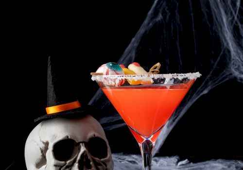 A red cocktail in a martini glass, topped with candy, sits beside a skull wearing a witch's hat, amidst Halloween decorations like spider webs.