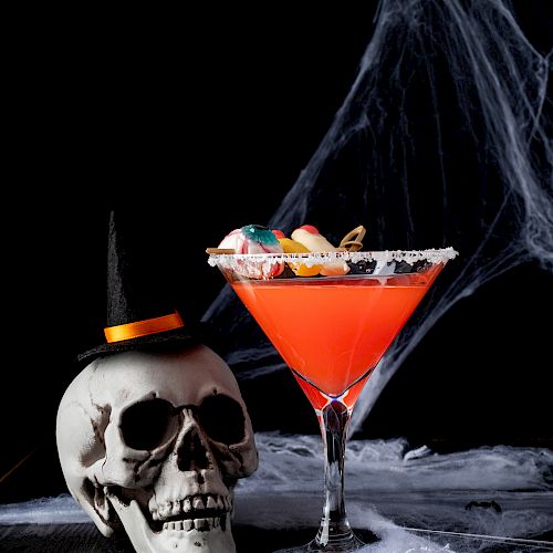 A red cocktail in a martini glass, topped with candy, sits beside a skull wearing a witch's hat, amidst Halloween decorations like spider webs.