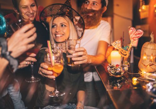 People in costumes are celebrating at a party, raising their glasses for a toast. Halloween decorations and festive lights adorn the scene.