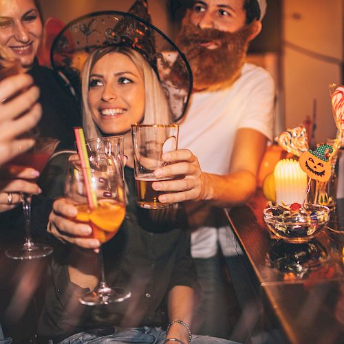 People in costumes are celebrating at a party, raising their glasses for a toast. Halloween decorations and festive lights adorn the scene.