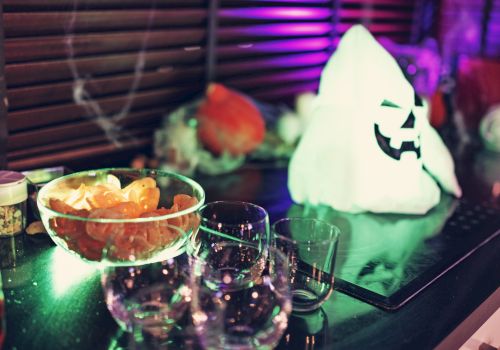 The image shows a festive setup with a glowing ghost decoration, a bowl of chips, several empty glasses, and additional party items in the background.