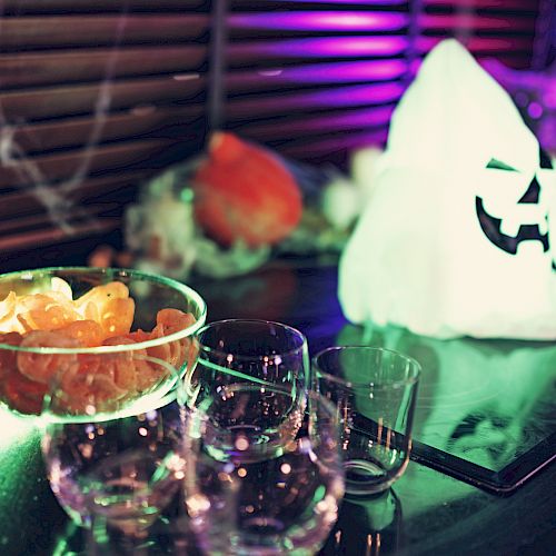 The image shows a festive setup with a glowing ghost decoration, a bowl of chips, several empty glasses, and additional party items in the background.