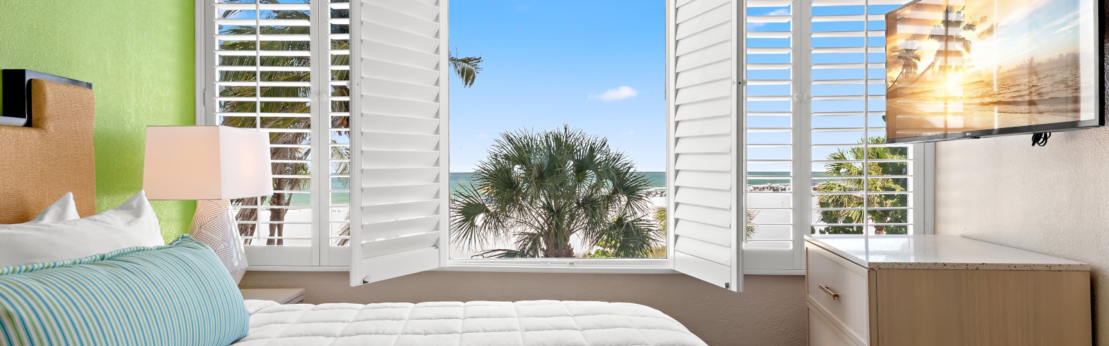 A bright bedroom with green and white walls, a bed with blue-striped pillow, open shutters revealing a palm tree view, and a wall-mounted TV.