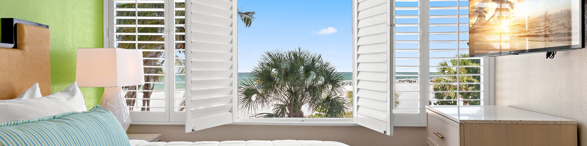 A bright bedroom with green and white walls, a bed with blue-striped pillow, open shutters revealing a palm tree view, and a wall-mounted TV.