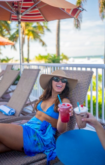 A woman in a sun hat and blue swimsuit relaxes on a lounge chair, holding a pink drink. Palm trees and sunny skies set the scene.