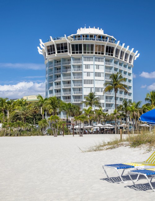 A beach scene with two lounge chairs and a blue umbrella under a clear sky, with a modern building and palm trees in the background.