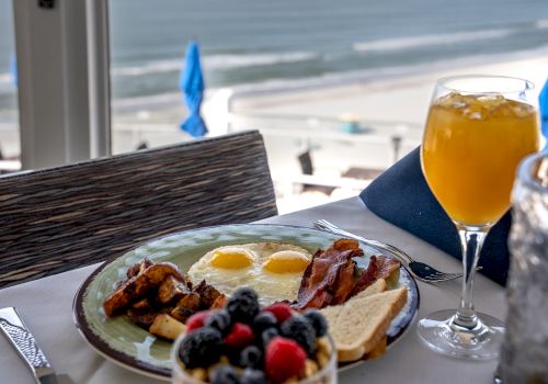 A breakfast setting with a plate of eggs, bacon, toast, and potatoes, a glass of orange juice, and a bowl of mixed berries by the beach, ends the sentence.