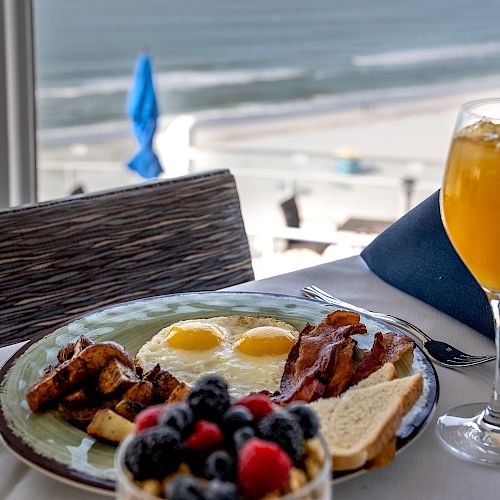 A breakfast setting with a plate of eggs, bacon, toast, and potatoes, a glass of orange juice, and a bowl of mixed berries by the beach, ends the sentence.