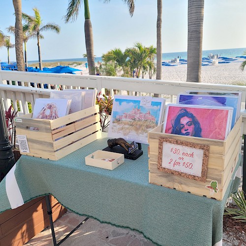 The image shows a seaside art display with wooden crates containing artwork, a table with a green cloth, and a sign indicating prices at $30 each.