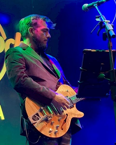 A bearded man is playing a guitar on stage with a microphone stand and a music stand in front of him, under colorful stage lights.