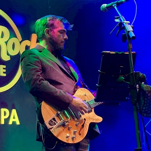 A bearded man is playing a guitar on stage with a microphone stand and a music stand in front of him, under colorful stage lights.