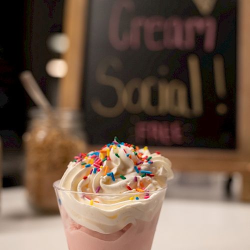 A cup of ice cream topped with whipped cream and sprinkles, with a blurred sign behind it saying 