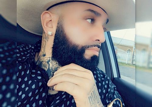 A bearded man with tattoos wearing a cowboy hat and patterned shirt, seated in a car looking out the window, with 