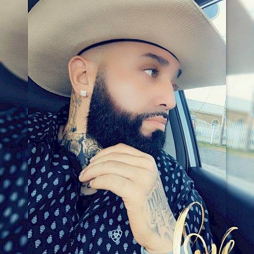 A bearded man with tattoos wearing a cowboy hat and patterned shirt, seated in a car looking out the window, with 