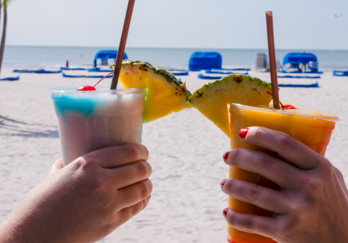 Two people toasting with tropical drinks garnished with pineapple slices. The scene is set on a beach with blue lounge chairs and palm trees.