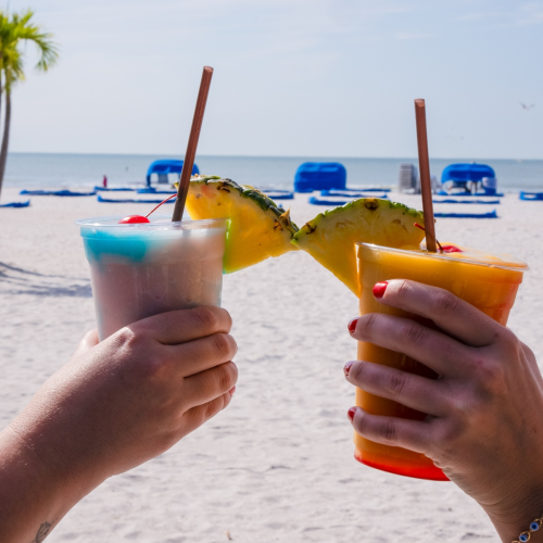 Two people toasting with tropical drinks garnished with pineapple slices. The scene is set on a beach with blue lounge chairs and palm trees.