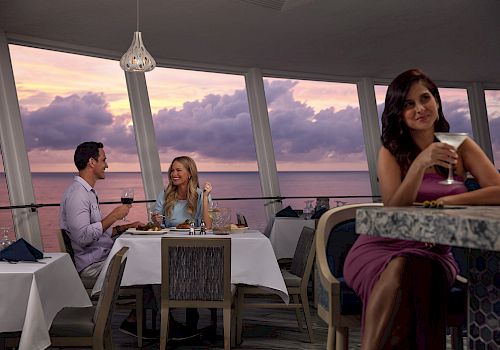 A couple is dining at a restaurant with scenic ocean views, while a woman in a purple dress enjoys a drink at the bar, all during sunset.