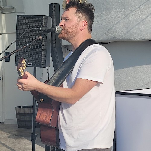 A man is playing a guitar and singing into a microphone. He is wearing a white T-shirt and standing in front of a speaker in an indoor setting.