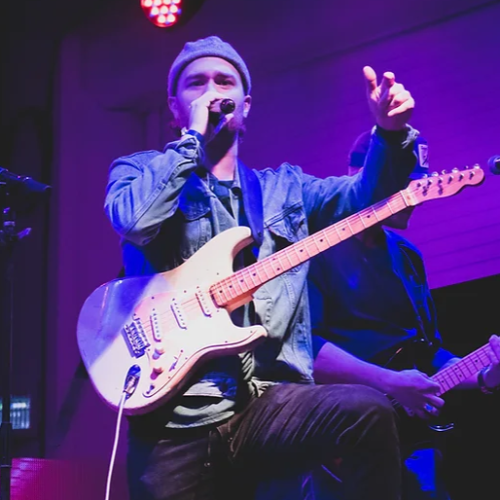 A musician wearing a beanie and denim jacket sings into a microphone while playing an electric guitar on stage under purple lighting.