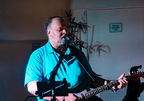 A man in a blue shirt is playing an electric guitar and singing into a microphone in a dimly lit room.