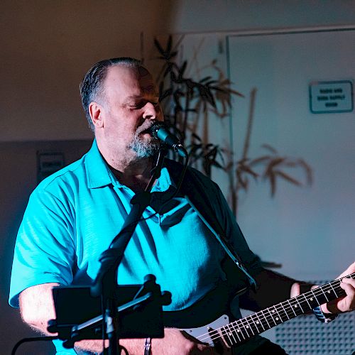 A man in a blue shirt is playing an electric guitar and singing into a microphone in a dimly lit room.