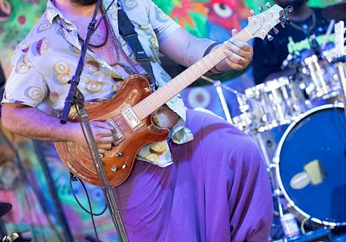 A bearded man with sunglasses playing an electric guitar on stage, with a colorful, animated mural in the background.