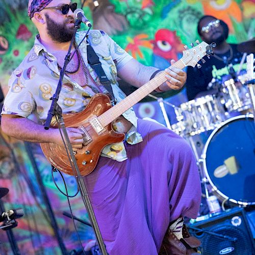 A bearded man with sunglasses playing an electric guitar on stage, with a colorful, animated mural in the background.