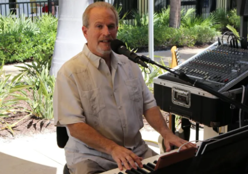 A man is playing a keyboard and singing into a microphone outdoors near a sound mixer, with greenery and a building in the background.