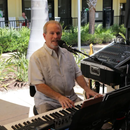 A man is playing a keyboard and singing into a microphone outdoors near a sound mixer, with greenery and a building in the background.