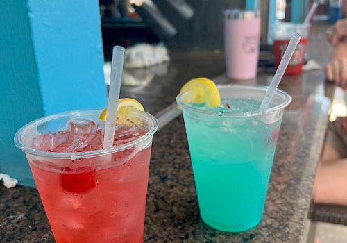 Two colorful cocktails, one red with cherries and one blue with a lemon slice, sit on a bar counter with a blurred background.