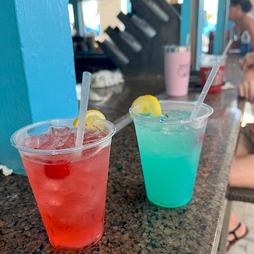 Two colorful cocktails, one red with cherries and one blue with a lemon slice, sit on a bar counter with a blurred background.