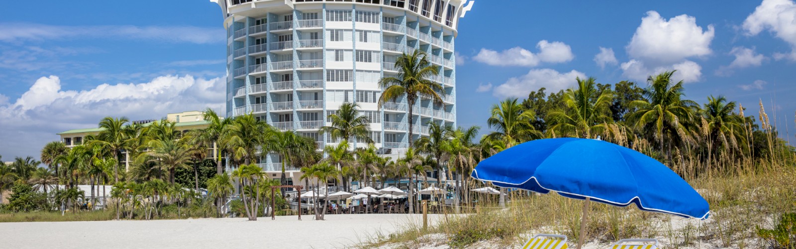 A tall, modern building is situated behind a sandy beach with palm trees, beach chairs, and a blue umbrella under a clear, blue sky.