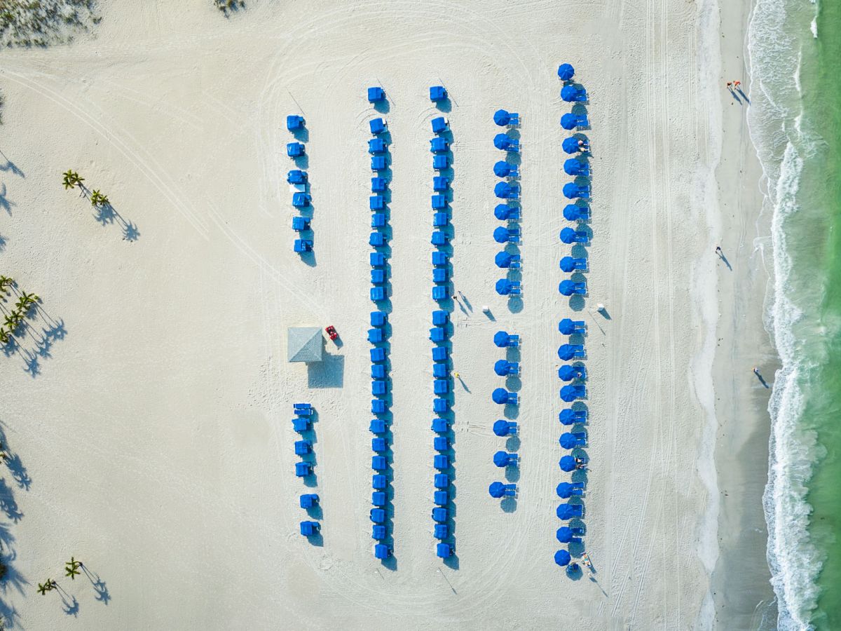 An aerial view of a beach with blue umbrellas neatly arranged in rows alongside the shoreline with a few people walking.