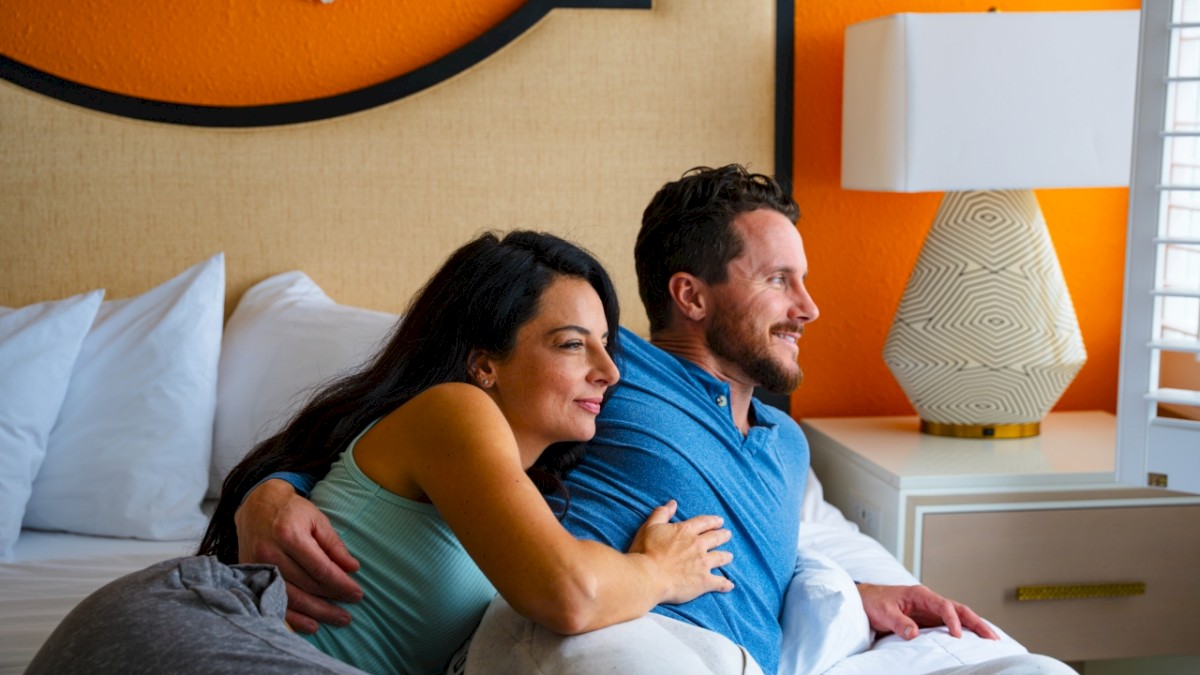 A couple is sitting on a bed, cuddling and smiling, while looking out a window in a brightly lit room with an orange wall and a table lamp.