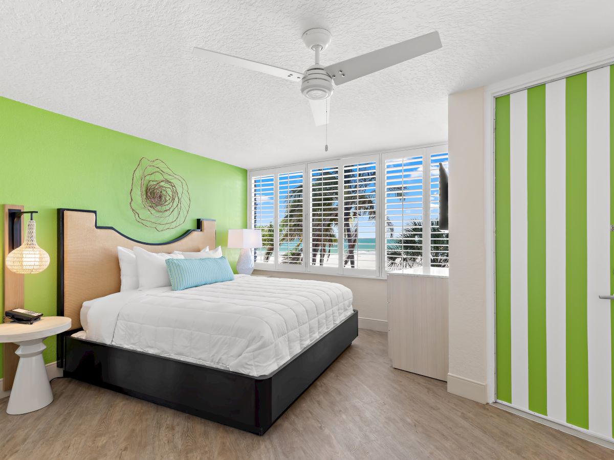 A modern bedroom with a green-accented wall, featuring a white bed, minimalist decor, a ceiling fan, and a window with a view of palm trees and the ocean.