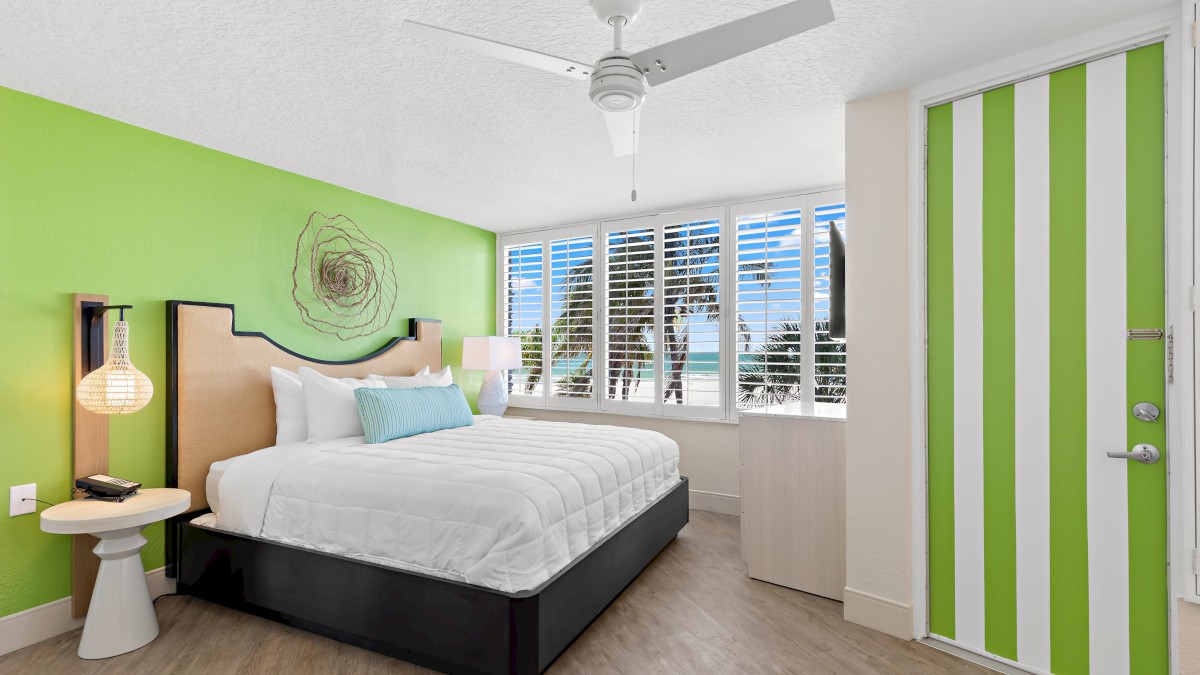 A modern bedroom with a green-accented wall, featuring a white bed, minimalist decor, a ceiling fan, and a window with a view of palm trees and the ocean.