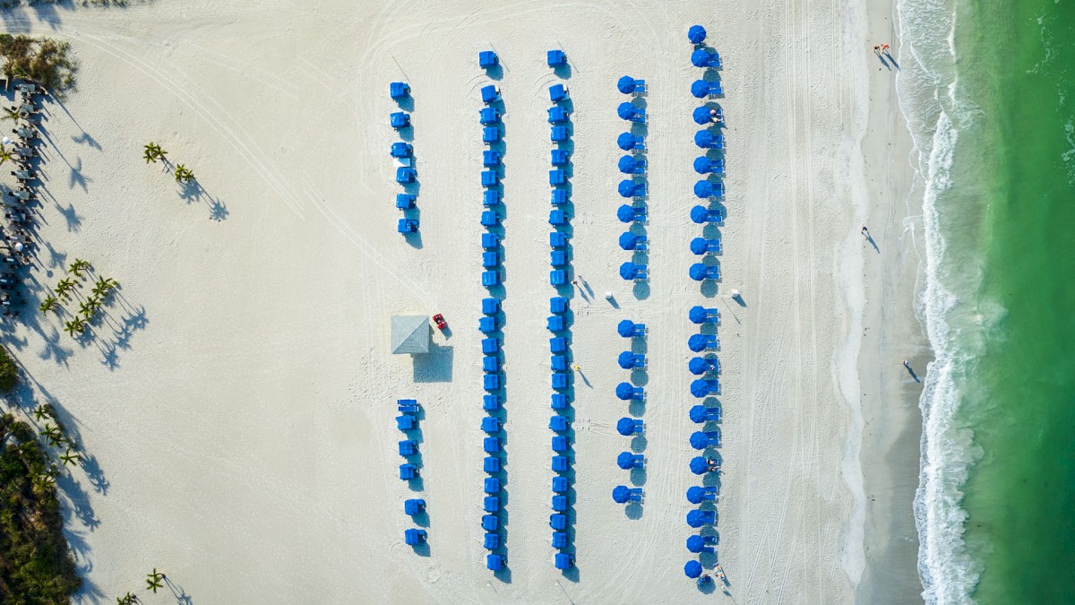 An aerial view of a beach with blue umbrellas neatly arranged in rows alongside the shoreline with a few people walking.