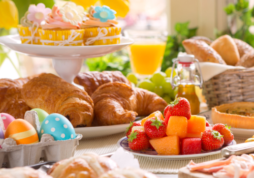 The image features a festive breakfast spread with croissants, colorful decorated eggs, fruit, pastries, juice, and flowers in the background.